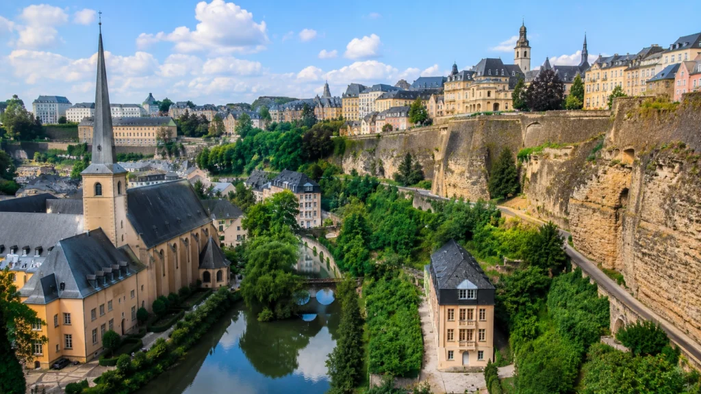 Luxembourg City’s historic lower town with a river, church spire, stone bridges, fortress cliffs, and colorful buildings.