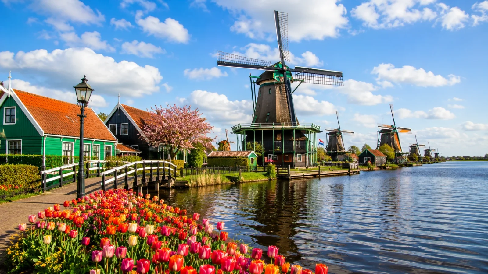 Traditional Dutch windmills beside a canal with colorful tulips, wooden houses, and a bright blue sky.