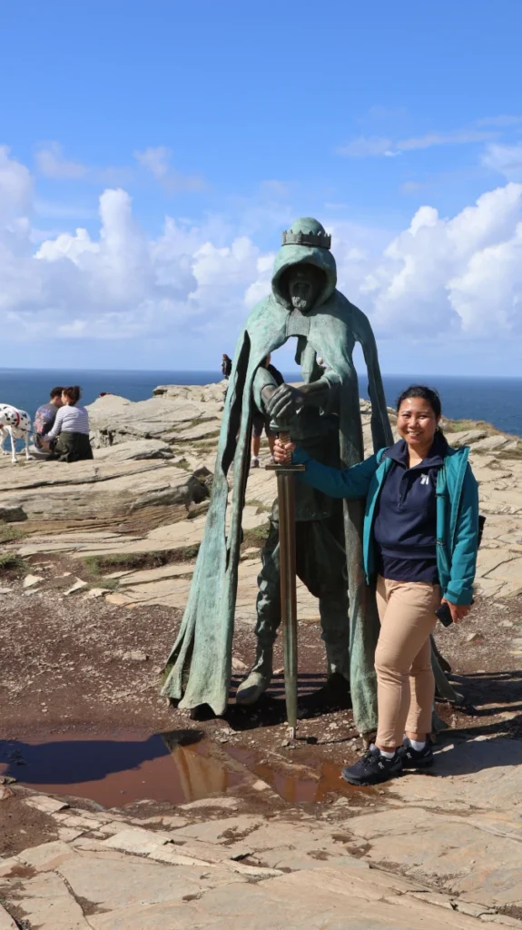 Visitor standing next to the Gallos statue at Tintagel Castle with the Cornish coastline behind