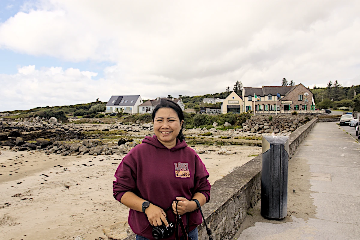 Woman standing at Furbo Beach near Galway with the rocky shoreline and coastal buildings in the background