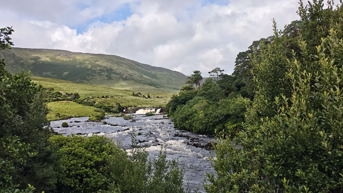 Aasleagh Falls on the River Erriff in County Mayo, Ireland, framed by trees with green hills and a cloudy sky behind