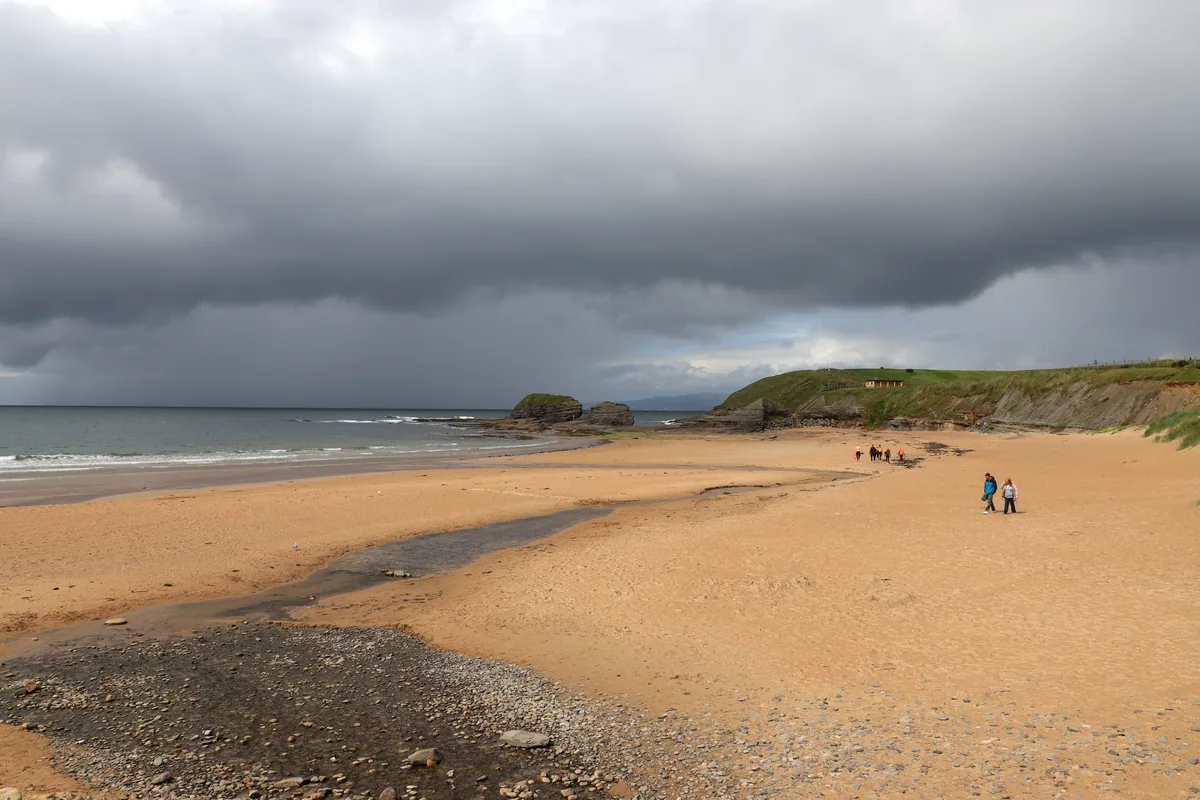 Wide sandy Bundoran Beach in County Donegal, Ireland, with a stream across the sand, a few walkers and dark rain clouds over the sea