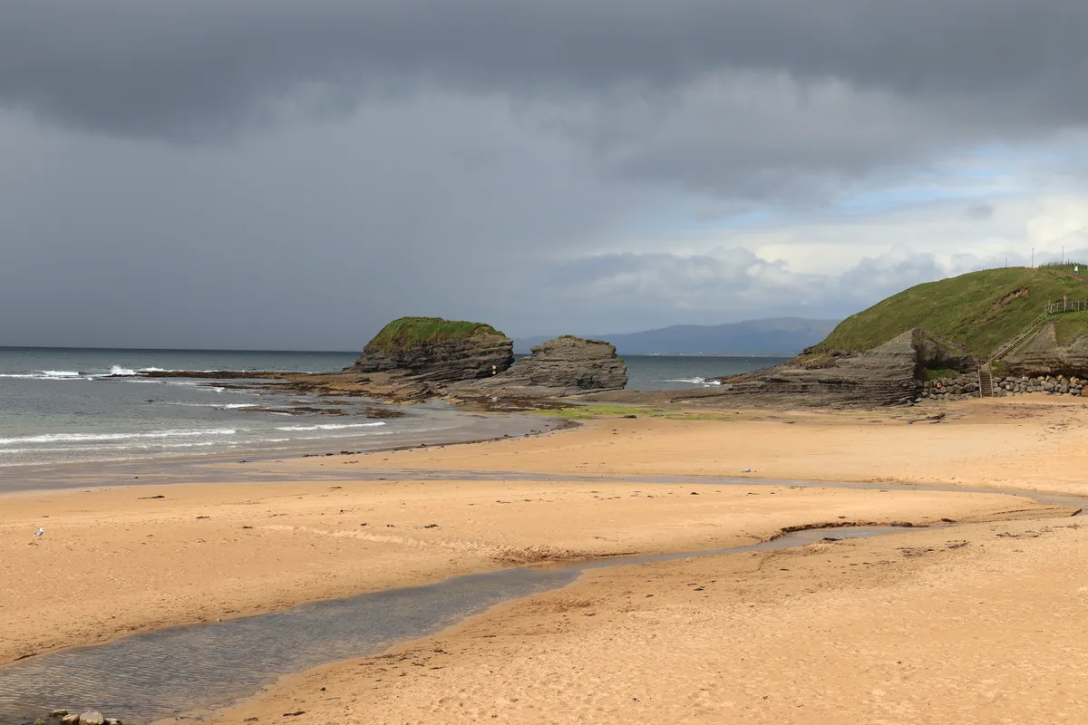 Golden sand, layered sea stacks and a grassy headland at Bundoran Beach, County Donegal, Ireland, with mountains across the bay