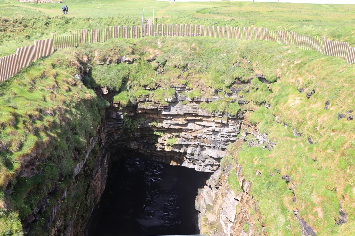 Downpatrick Head blowhole in County Mayo, Ireland: a deep opening in the grass with layered rock walls, dark water below and a fence around it.