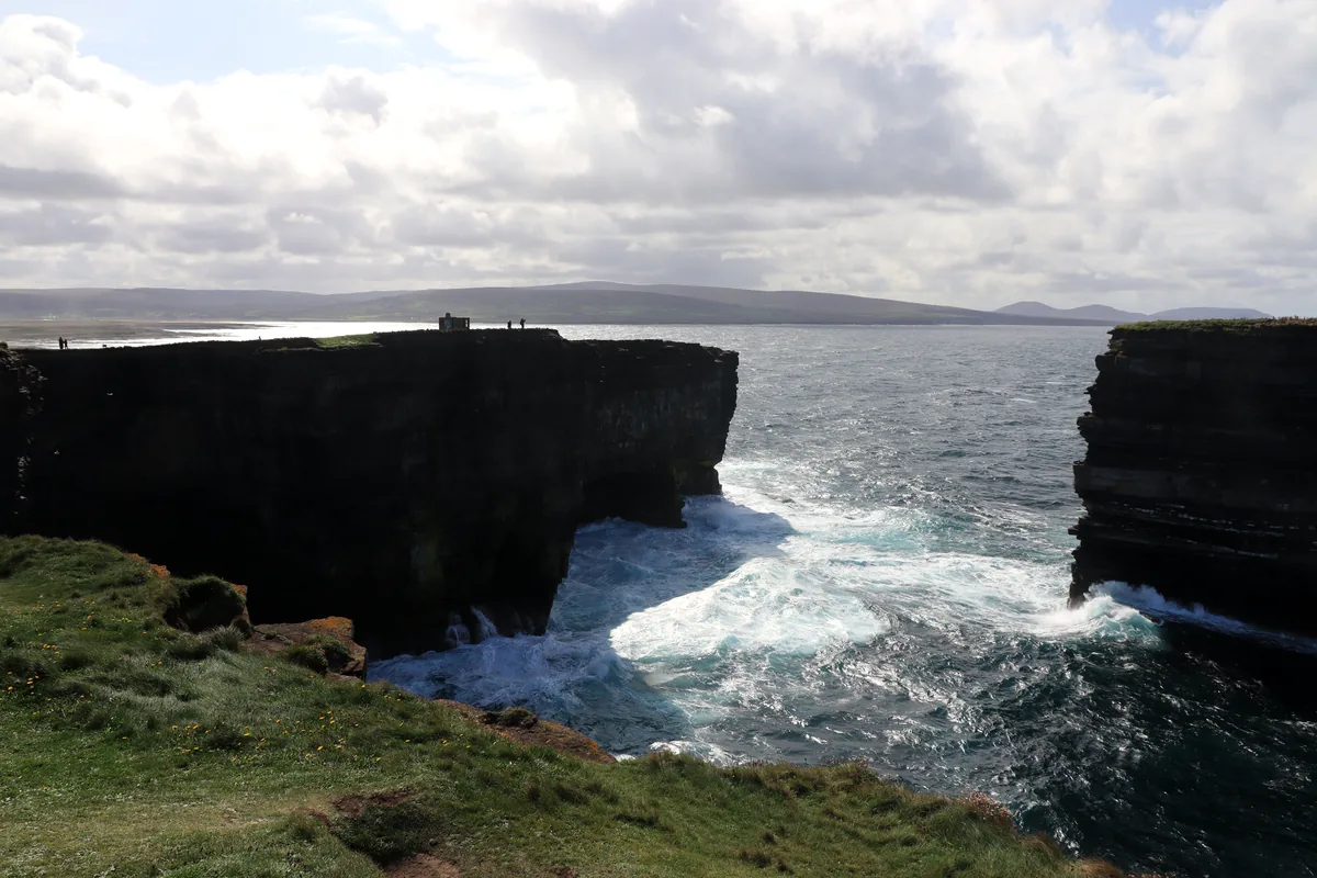 Downpatrick Head cliffs in County Mayo, Ireland, with surf between the headland and the Dun Briste sea stack and small figures on the clifftop.