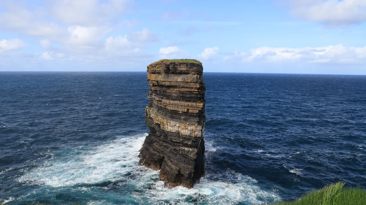 Dun Briste sea stack at Downpatrick Head, County Mayo, with layered rock, a grass top and waves breaking around its base.