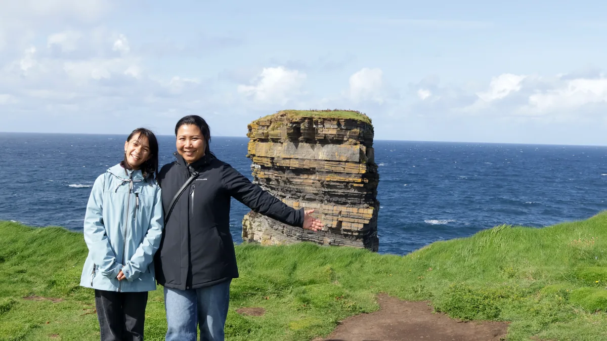Two members of THE OVRTLND team smile on the grass at Downpatrick Head, County Mayo, with the Dun Briste sea stack behind them.