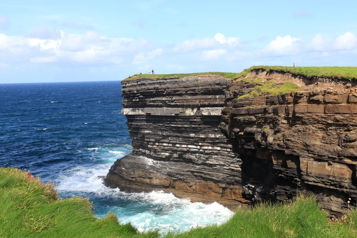 Downpatrick Head cliff edge in County Mayo, Ireland: visitors stand on the grass above a sheer layered rock face with no fence.