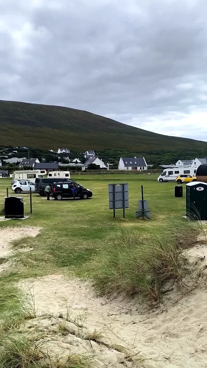 Unsurfaced grass car park at Dugort Beach, Achill Island, with campervans, a portable toilet and bins below Slievemore