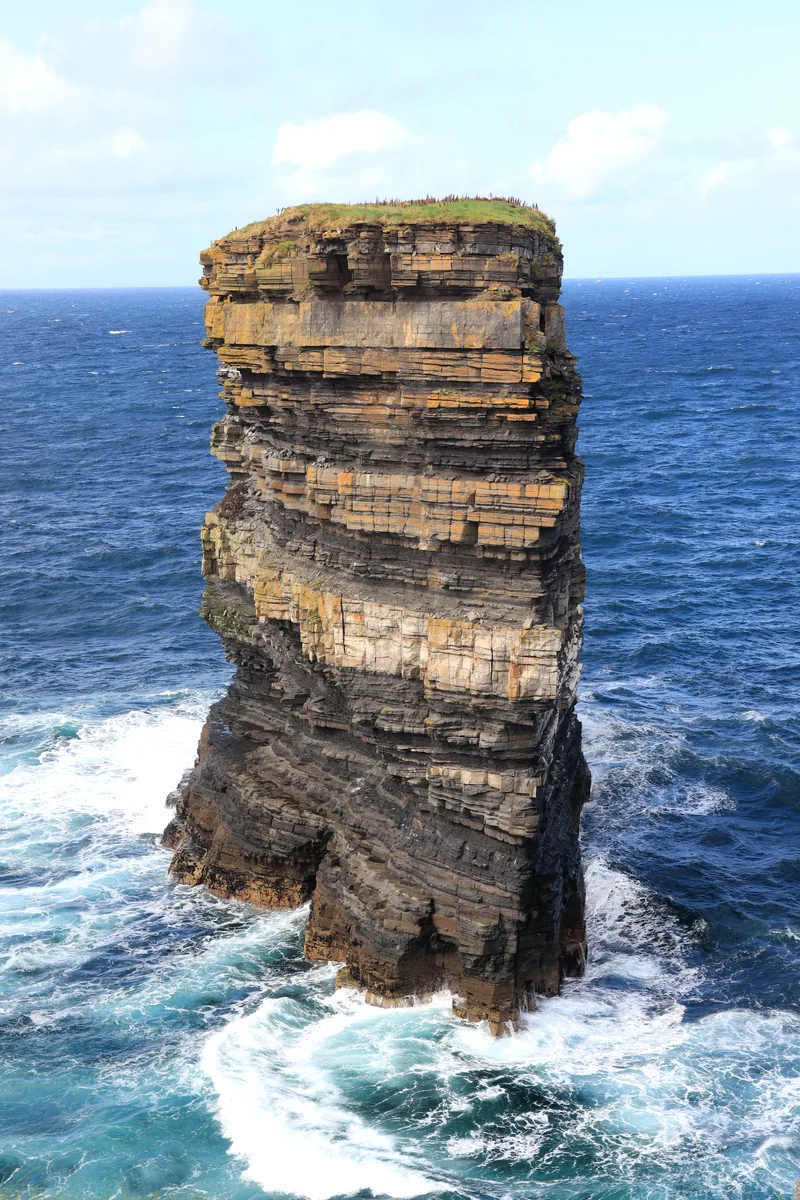 Dun Briste sea stack at Downpatrick Head, County Mayo, seen close up with bands of dark and yellow rock and white surf at its base.