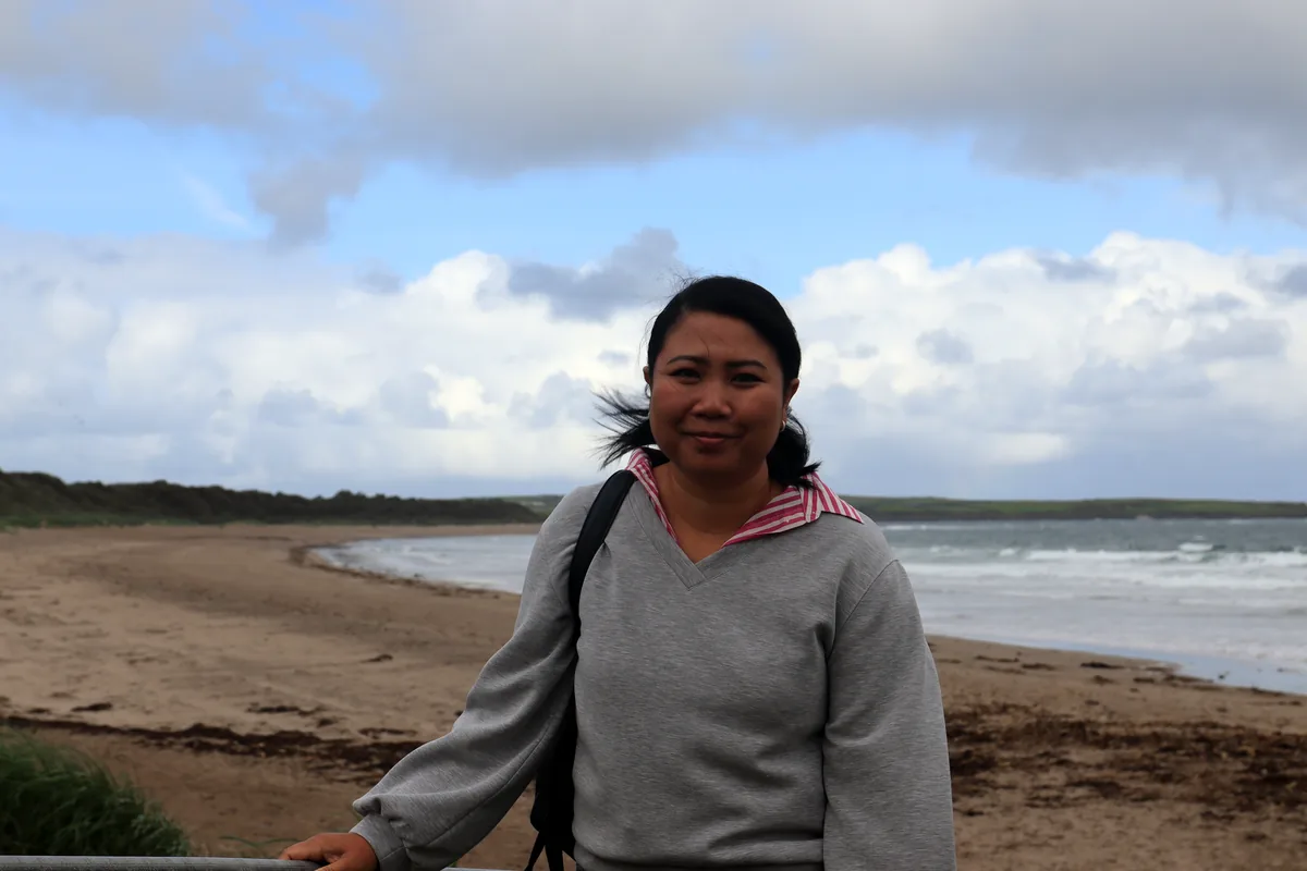 A visitor stands at the railing above Dunmoran Beach in County Sligo, Ireland, with wind in her hair and waves rolling onto the sand.
