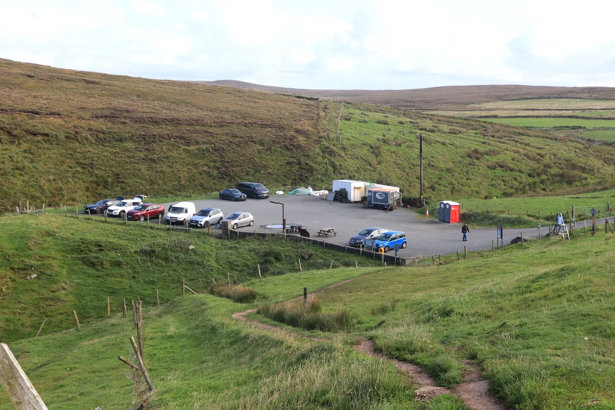 Erris Head car park in County Mayo, Ireland, seen from the path, with parked cars, picnic tables and two portable toilets.