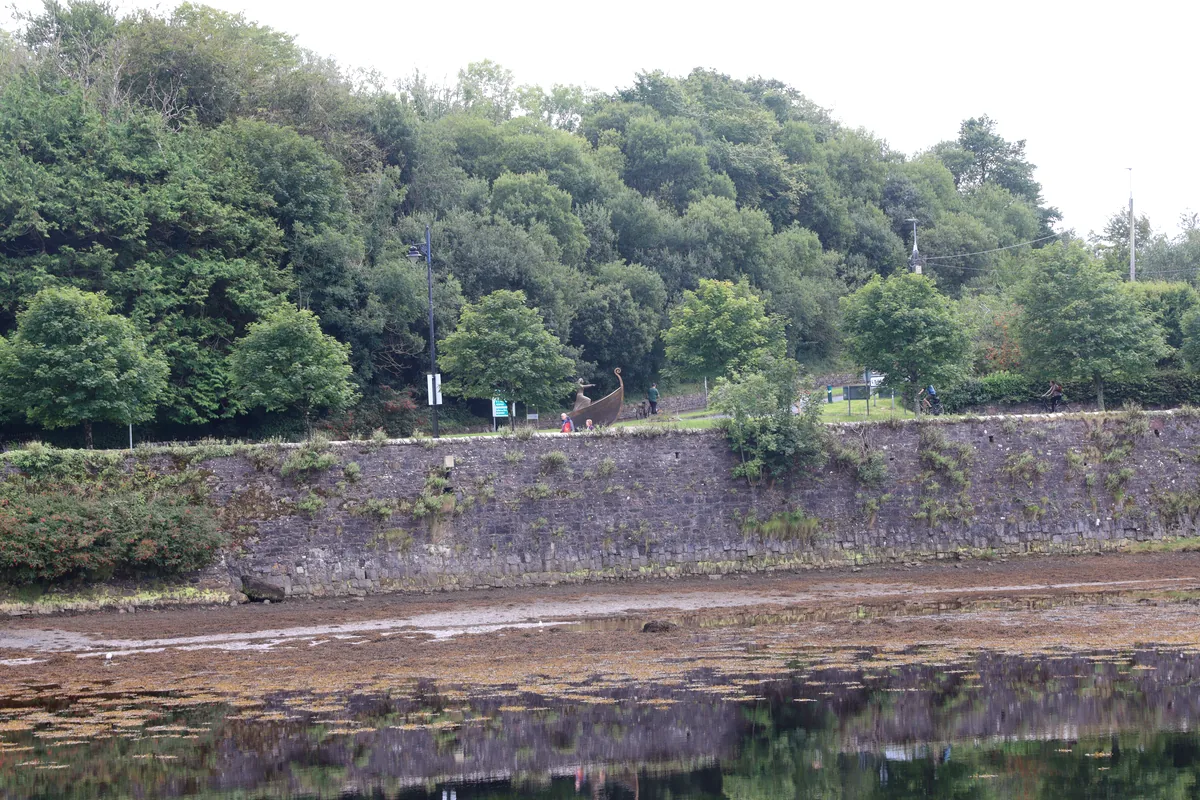 Newport Mayo stone river wall at low tide, with a ship sculpture, walkers and cyclists on the path above, County Mayo, Ireland
