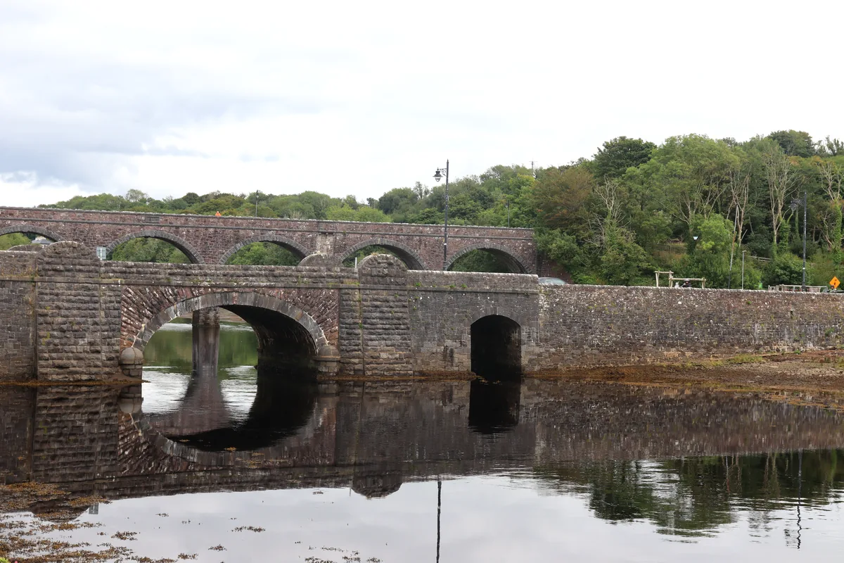 Newport Mayo stone road bridge with the red sandstone Seven Arches railway viaduct behind, both reflected in the Black Oak River, County Mayo