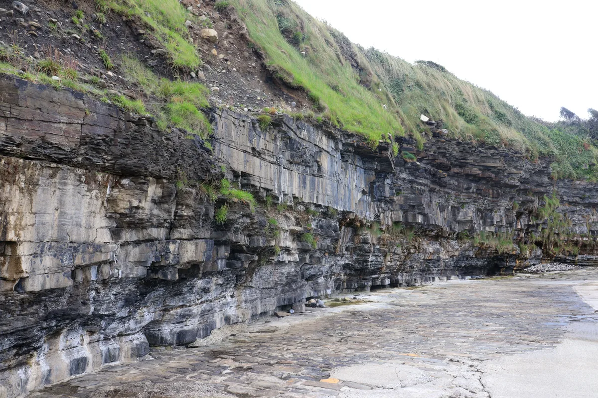 Layered rock cliff topped with grass beside the slipway at Pollnadivva Pier in County Sligo, Ireland.