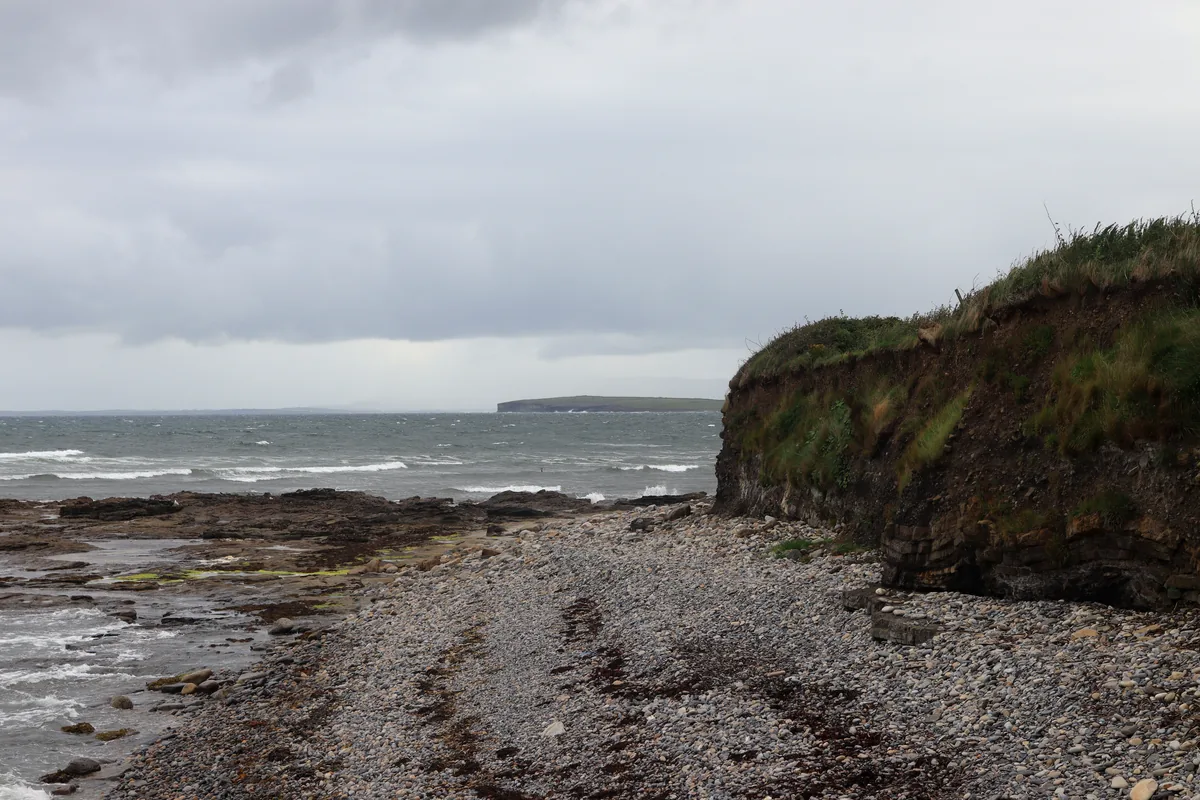 Pebble shore and low grassy cliff next to Pollnadivva Pier in Co. Sligo, with waves and a flat headland on the horizon under grey clouds.