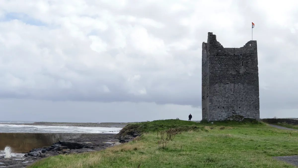 Roslee Castle, a ruined stone tower with an Irish flag, standing on grass beside Easkey pier under a cloudy sky in County Sligo.