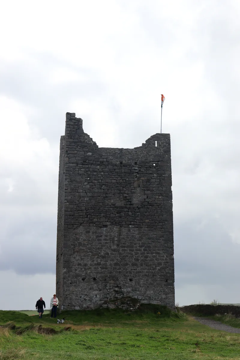 Roslee Castle, a tall ruined stone tower with an Irish flag on top, in Easkey, County Sligo, with two visitors and a dog walking past.