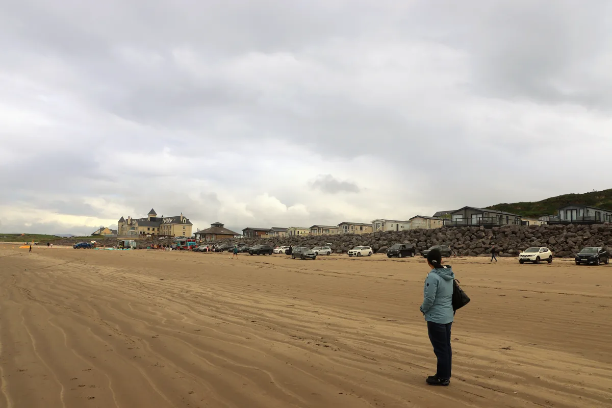 A visitor on Rossnowlagh Beach looking at cars parked on the sand in front of the rock wall and holiday homes in Donegal