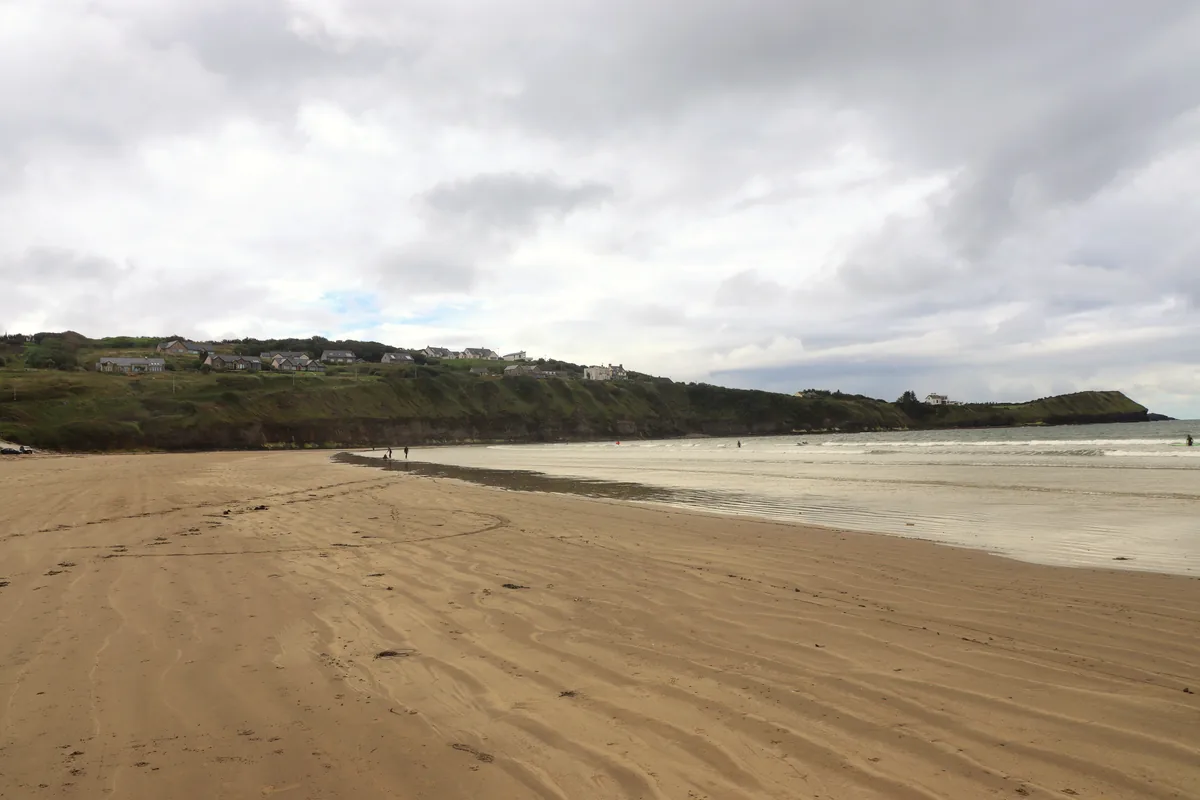 Wet rippled sand and gentle surf at Rossnowlagh Beach in Donegal, with surfers in the water and houses on the green cliffs above